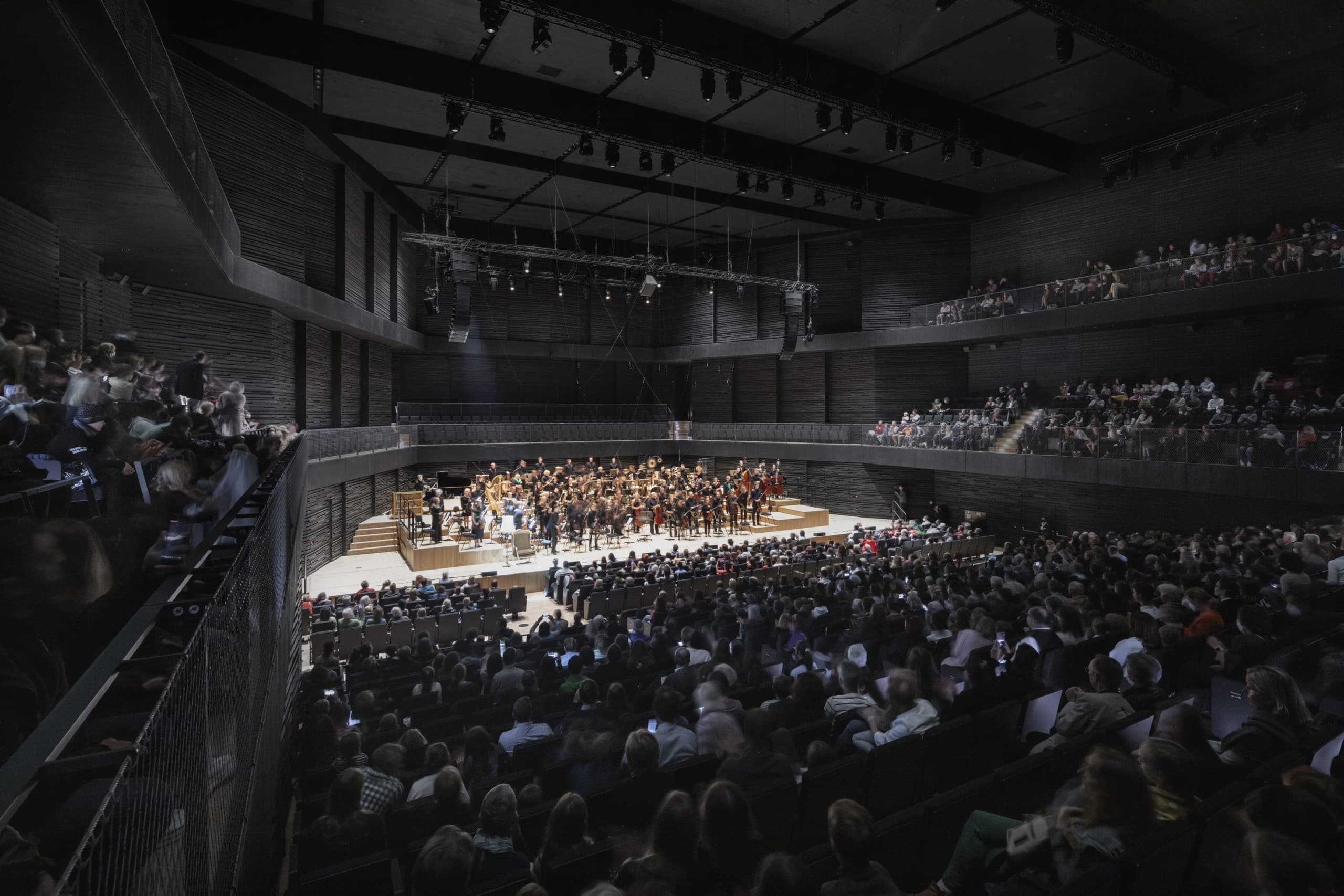 Blick in den vollbesetzten Konzertsaal der Isarphilharmonie München. Dunkel die Wandungen, Spot auf das Orchester. Foto: HG Esch, © & Courtesy gmp.